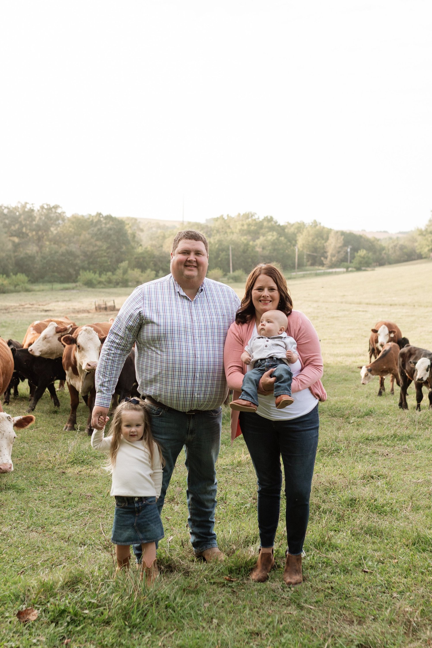 Family portrait on a farm with cows in the background, featuring a man and woman holding their baby, while a young girl stands beside them. The setting showcases a lush green field and trees, highlighting a rural lifestyle.