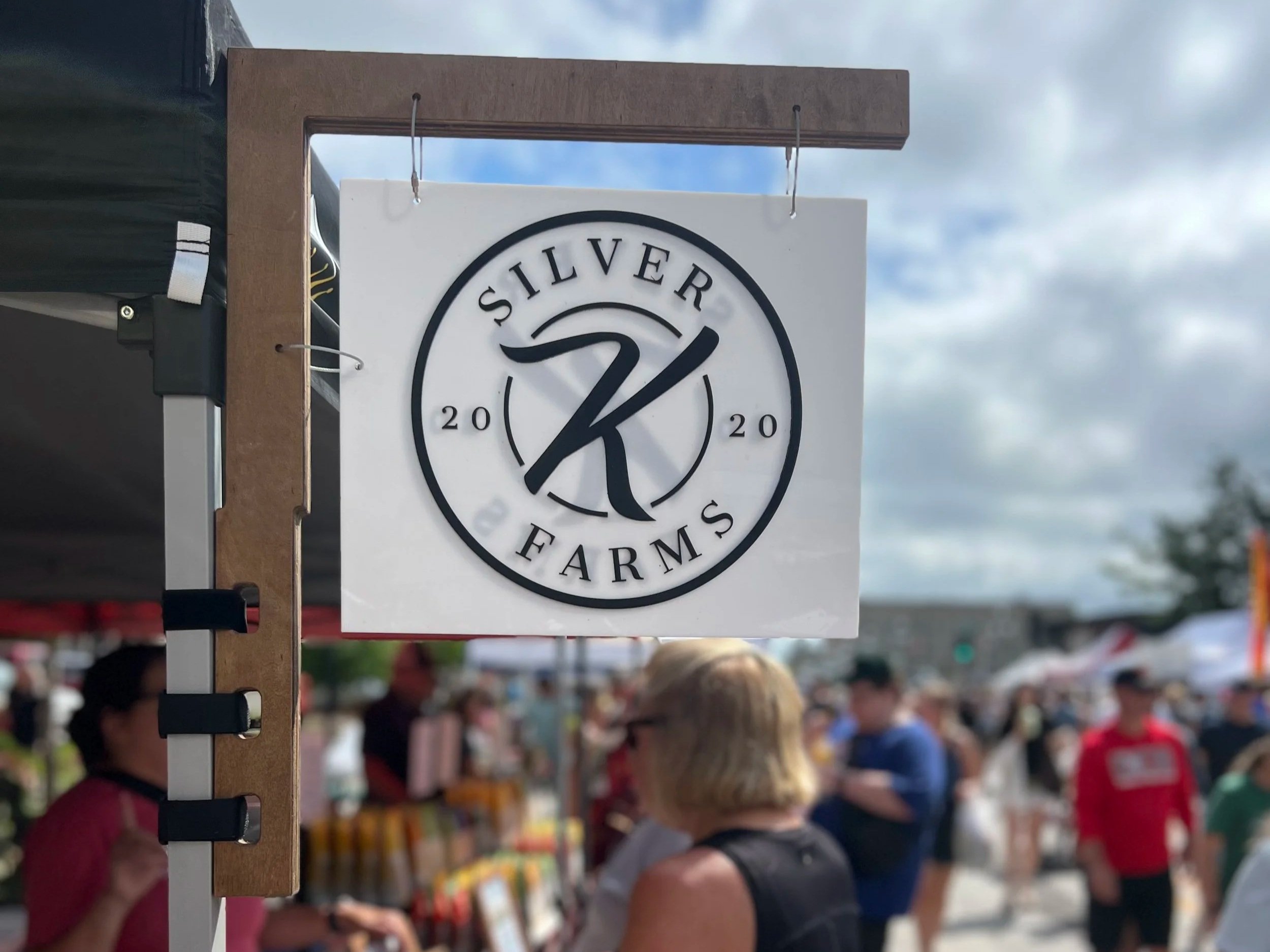 Sign for Silver Farms displayed at a bustling outdoor market, showcasing the brand logo and the year 2020, with a crowd of visitors in the background.