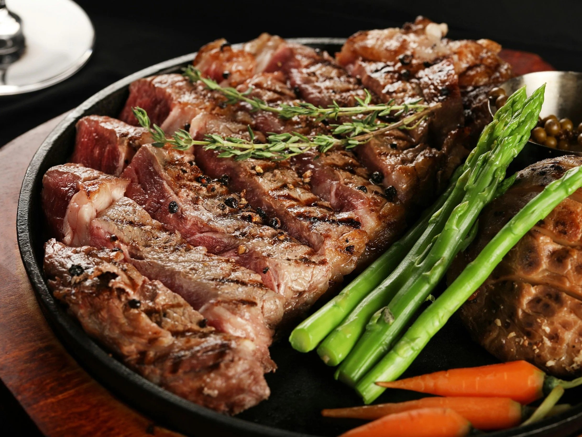 Juicy grilled steak slices garnished with fresh thyme, served alongside vibrant asparagus and baby carrots on a sizzling platter. Perfect for meat lovers and gourmet dining experiences.